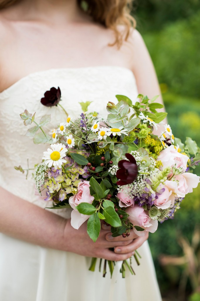 Bridal bouquet of white, pink and purple flowers, held by a woman wearing a white dress