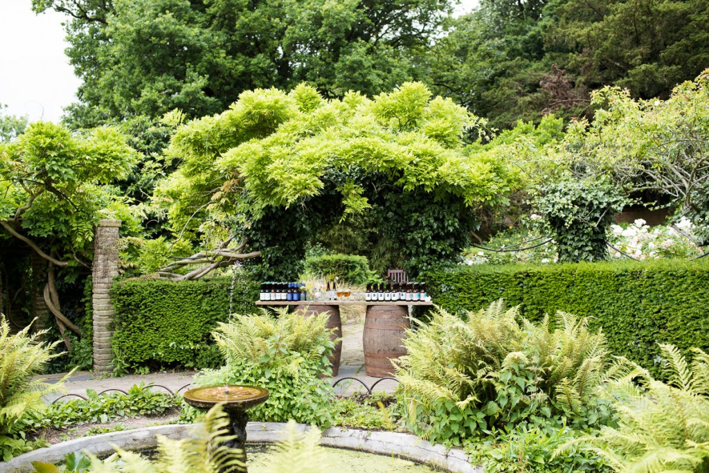 A garden with a fountain in the foreground and a tabletop in the background arranged with beer bottles